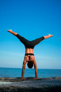 Full length of woman exercising at beach against clear blue sky