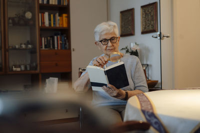 Senior woman reading book with magnifying glass at home