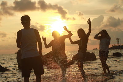 Friends splashing water on shore at beach against sky during sunset