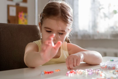 A young girl with pigtails creates colorful jewelry by stringing beads at a well-lit table. 
