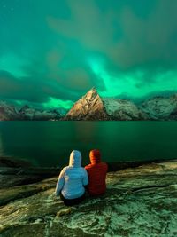 Rear view of hikers sitting on rock by lake against aurora