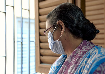 An aged bengali woman wearing surgical face mask, casually looking out of window