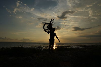 Silhouette person holding bicycle on beach against sky during sunset