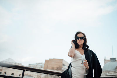 Low angle view of woman wearing sunglasses and champagne flute standing by railing against sky