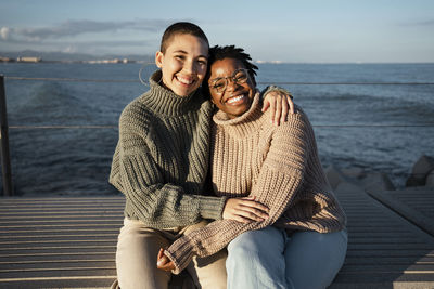 Happy woman sitting with arm around on friend against sky