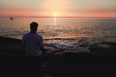 Rear view of man sitting on rock against sea during sunset