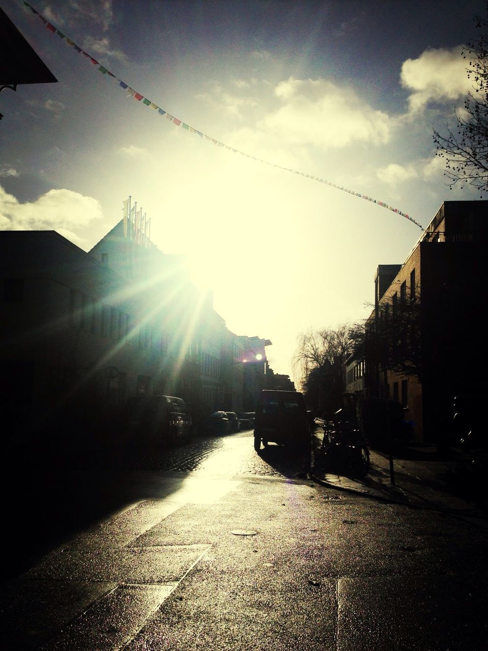 building exterior, architecture, built structure, sun, sky, sunlight, sunbeam, transportation, street, city, sunset, lens flare, road, the way forward, car, cloud - sky, silhouette, building, cloud, residential structure