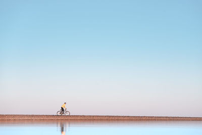 Man riding bicycle on riverbank against clear sky