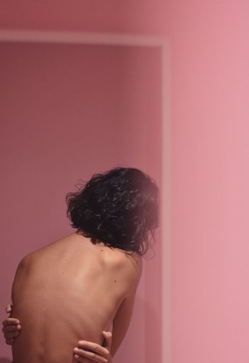 Rear view of shirtless sensuous woman standing against pink wall