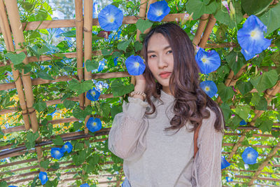 Portrait of young woman holding flower while standing against morning glory flower plant