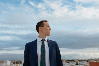 Young businessman in a suit and tie looking away thoughtful and smiling outside with a cloudy sky