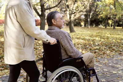 Disabled senior man sitting in wheelchair pushed by woman at park