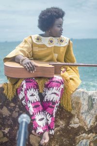 Full length of woman sitting on rock with guitar against sea