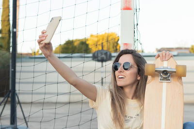 Young caucasian girl laughing while taking a selfie sitting next to a net with her longboard