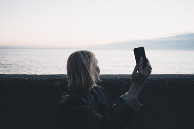 Woman taking selfie through mobile phone while standing by lake during sunset