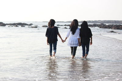 Rear view of friends holding hands while walking in sea on shore at beach