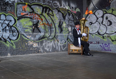 Man playing tuba while sitting against graffiti wall