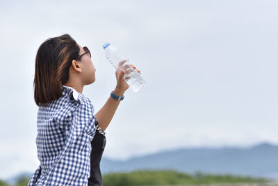 Side view of woman drinking water from bottle against sky