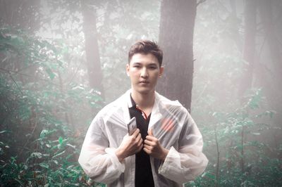 Portrait of man wearing raincoat while standing in forest during rainy season