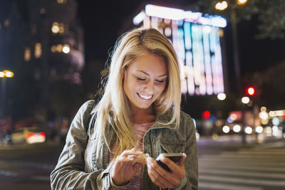 Happy young woman using smart phone on city street at night