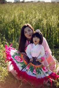 Korean woman with a daughter sits on green field in summer holding fan sunset in national costumes