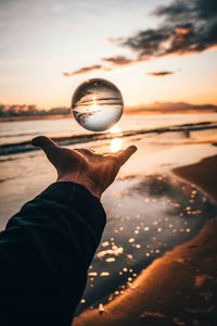 Cropped hand of person catching crystal ball against sky during sunset