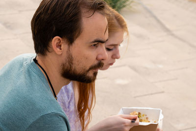 Focused young couple eating takeaway food while sitting on stairs in street
