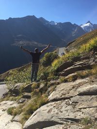 Rear view of man with arms outstretched standing on rock by road against mountains 