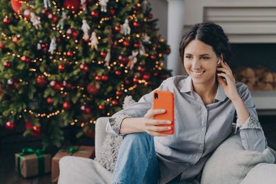 Young woman using mobile phone while sitting on christmas tree at home