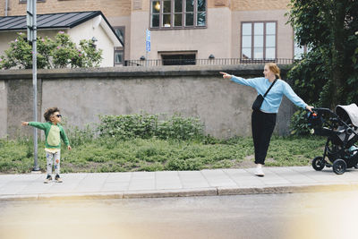 Boy pointing and showing mother with baby stroller while standing on sidewalk at city