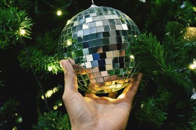 Close-up of hand holding disco ball on illuminated christmas tree at night