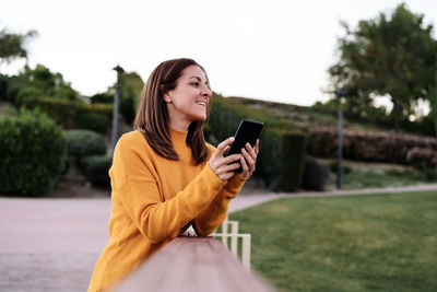 Smiling caucasian woman wearing yellow pullover using mobile phone outdoors in park during sunset. 