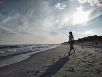 Silhouette woman walking on sand at beach against sky during sunny day