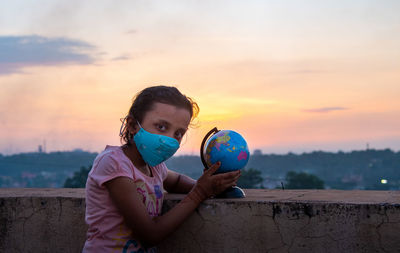 Portrait of girl holding man made globe against sky during sunset