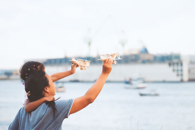 Rear view of girl and mother holding toy airplanes at beach against sky