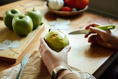 Halloween diy fruit ideas. female hands cutting out halloween green apple with creepy carved face