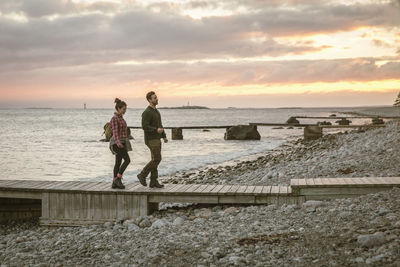 Side view of couple walking on jetty at beach against sky during sunset