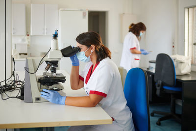 Female medical technologists in uniforms and sterile masks working with microscope in laboratory