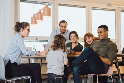 Smiling businessman showing smart phone to son while sitting by colleagues at creative office