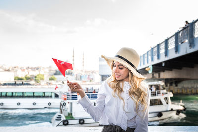 Young woman in hat holding flag while standing in boat against sky