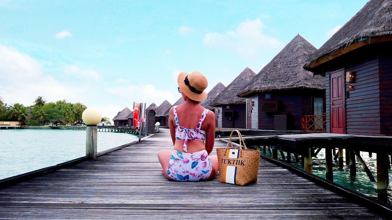 REAR VIEW OF WOMAN SITTING ON WOODEN STRUCTURE AGAINST SKY