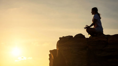 Low angle view of woman doing yoga on rock formation against sky during sunset