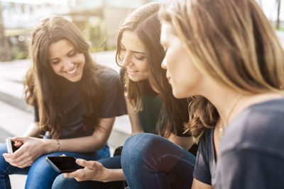 Three happy young women sitting outdoors looking at cell phone