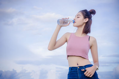 Woman drinking water from bottle while standing against sky