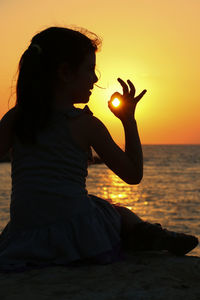 Rear view of girl sitting on beach against sky during sunset