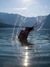 Woman tossing wet hair in lake against mountains during sunny day