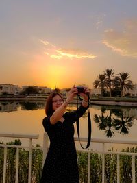 Young woman photographing with camera against sky during sunset