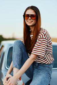 Portrait of young woman sitting on car against sky