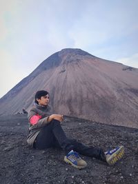 Full length of young man sitting on mountain against sky