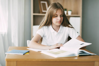 A pretty girl is reading a book sitting at a table, doing homework, preparing for exams. 